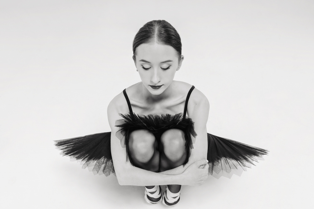 ein Porträt einer Ballerina in einem schwarzen Tutu,die hockt und ihre Knie umarmt von Alexandr